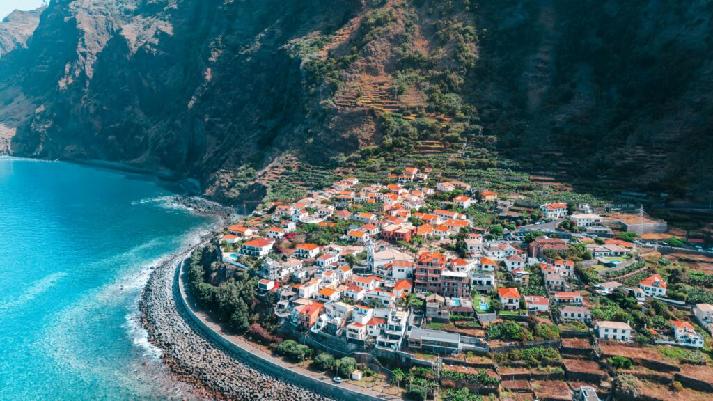 Island Hot List: European Summer Escapes for Every Vibe Stunning aerial view of a coastal town in Madeira, Portugal, with lush cliffs and turquoise sea.