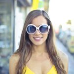 A stylish young woman with sunglasses smiles confidently on a sunny urban street.
